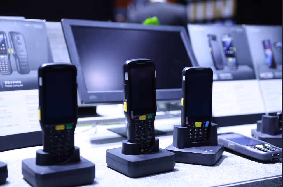 Three handheld barcode scanners on charging docks with a computer monitor and product information displays in the background.