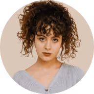 Portrait of a young woman with curly brown hair and blue eyes, wearing a light gray top against a beige background.