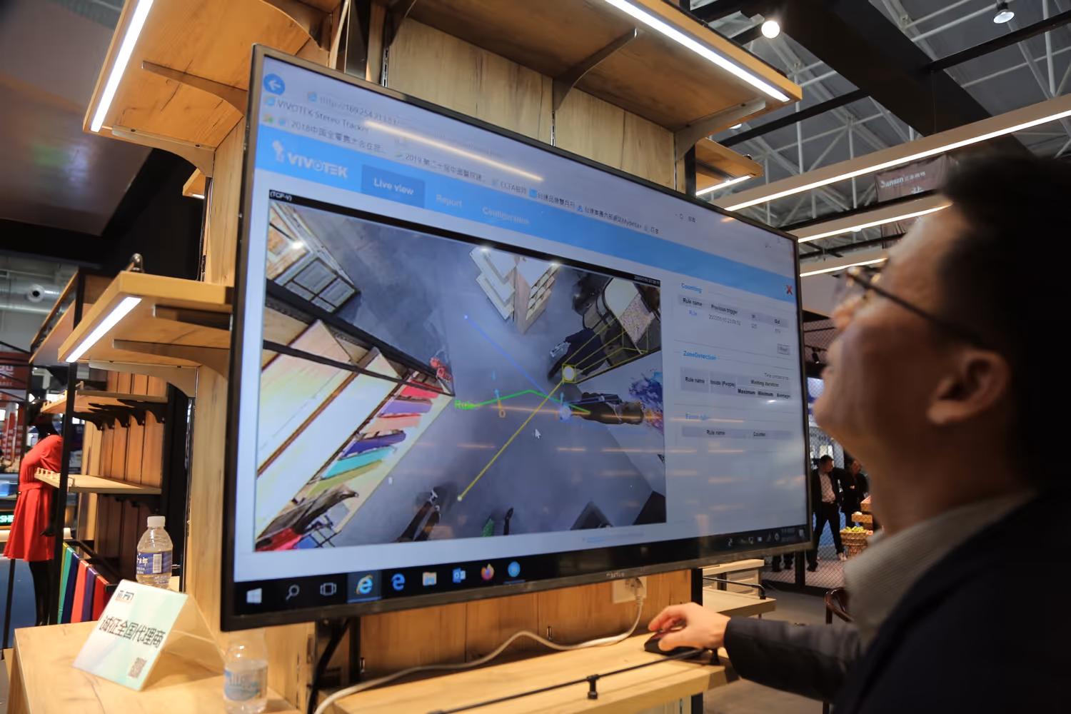 Man interacting with a large monitor displaying a surveillance system interface showing a top-down view of a store layout with colored zones and paths.