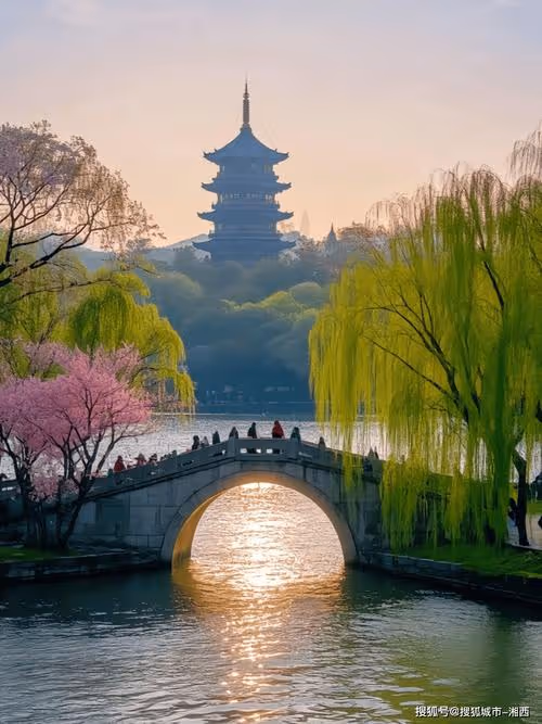 Stone arch bridge over a river with people walking, surrounded by green and pink blossoming trees, with a multi-tiered pagoda in the background at sunset.