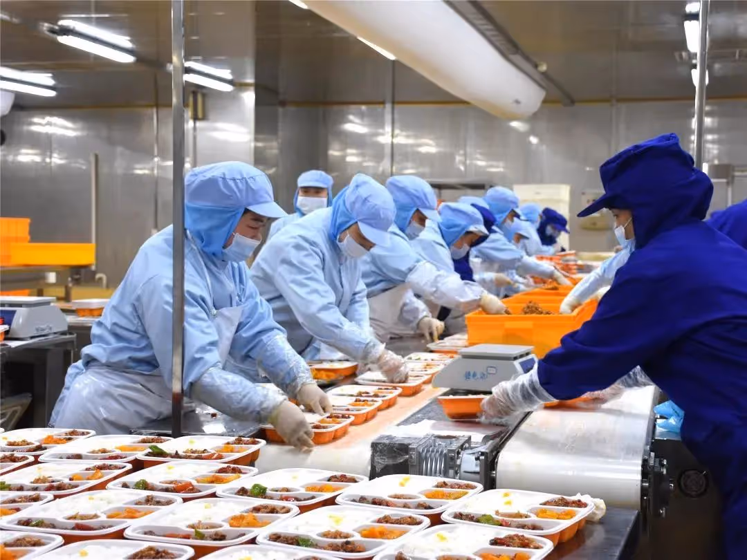Workers in blue protective clothing and face masks assembling and packaging meals on a production line in a food processing facility.