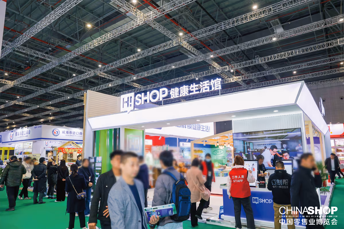 Busy exhibition booth with a lit sign reading 'H Shop' in Chinese, showcasing health-related products with several visitors and staff inside a large convention hall.