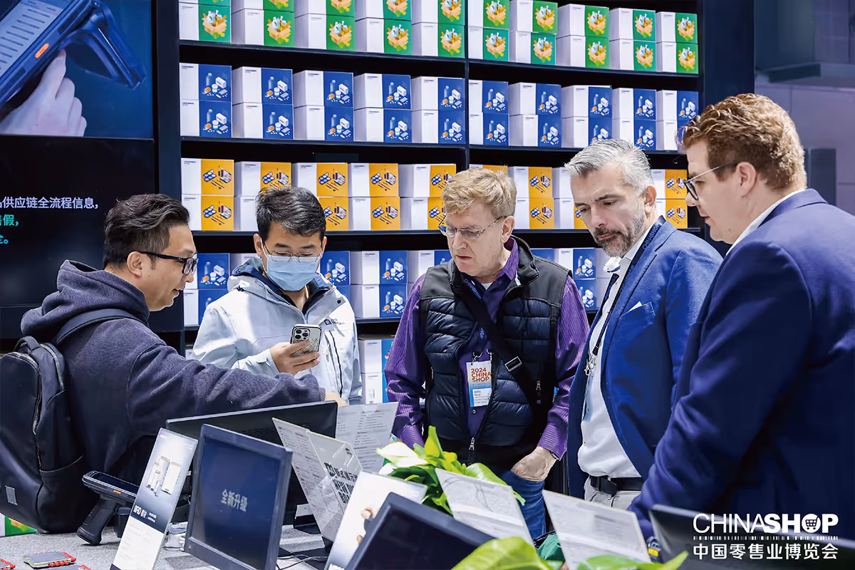 Five men examining products and information at a trade show booth with colorful boxes on shelves in the background and ChinaShop logo in the corner.