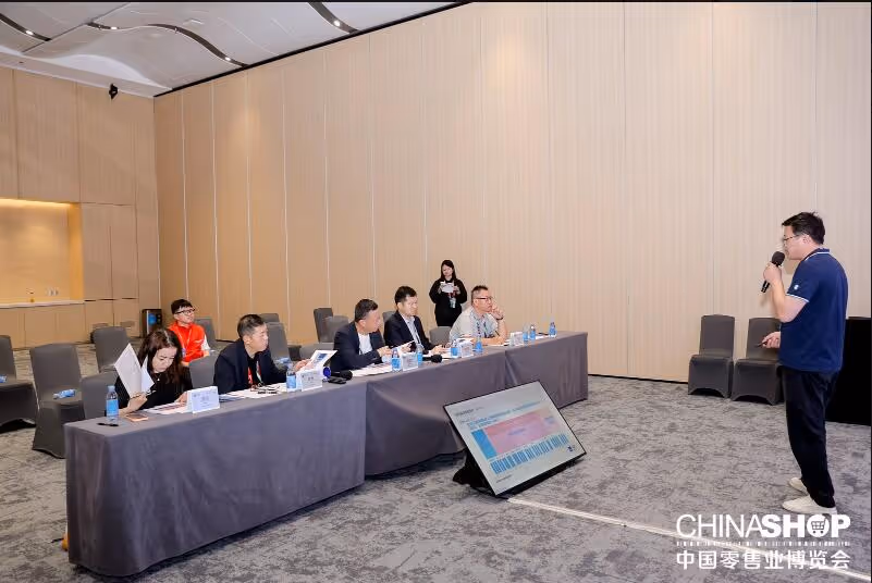 A man speaking into a microphone while six people seated at a long table listen and review documents in a conference room.
