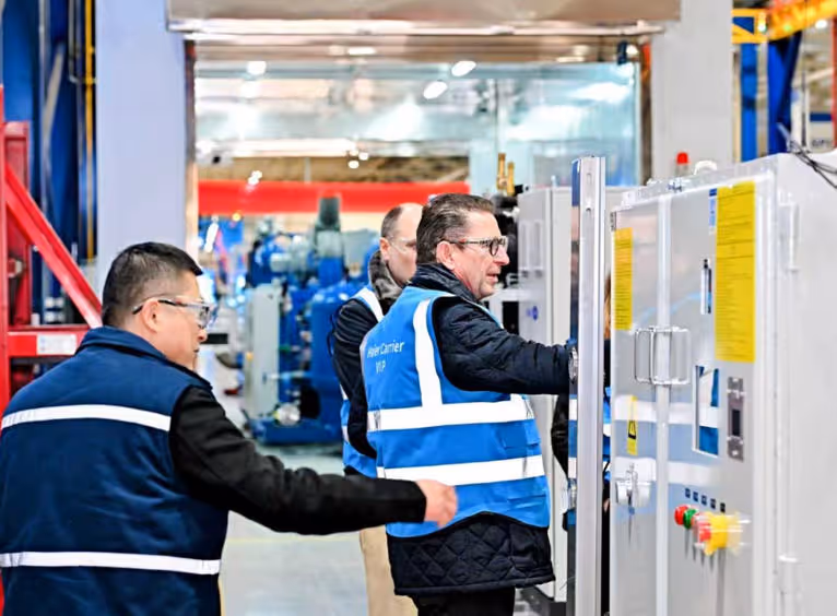 Three men wearing blue safety vests inspecting and operating industrial control panels inside a factory.