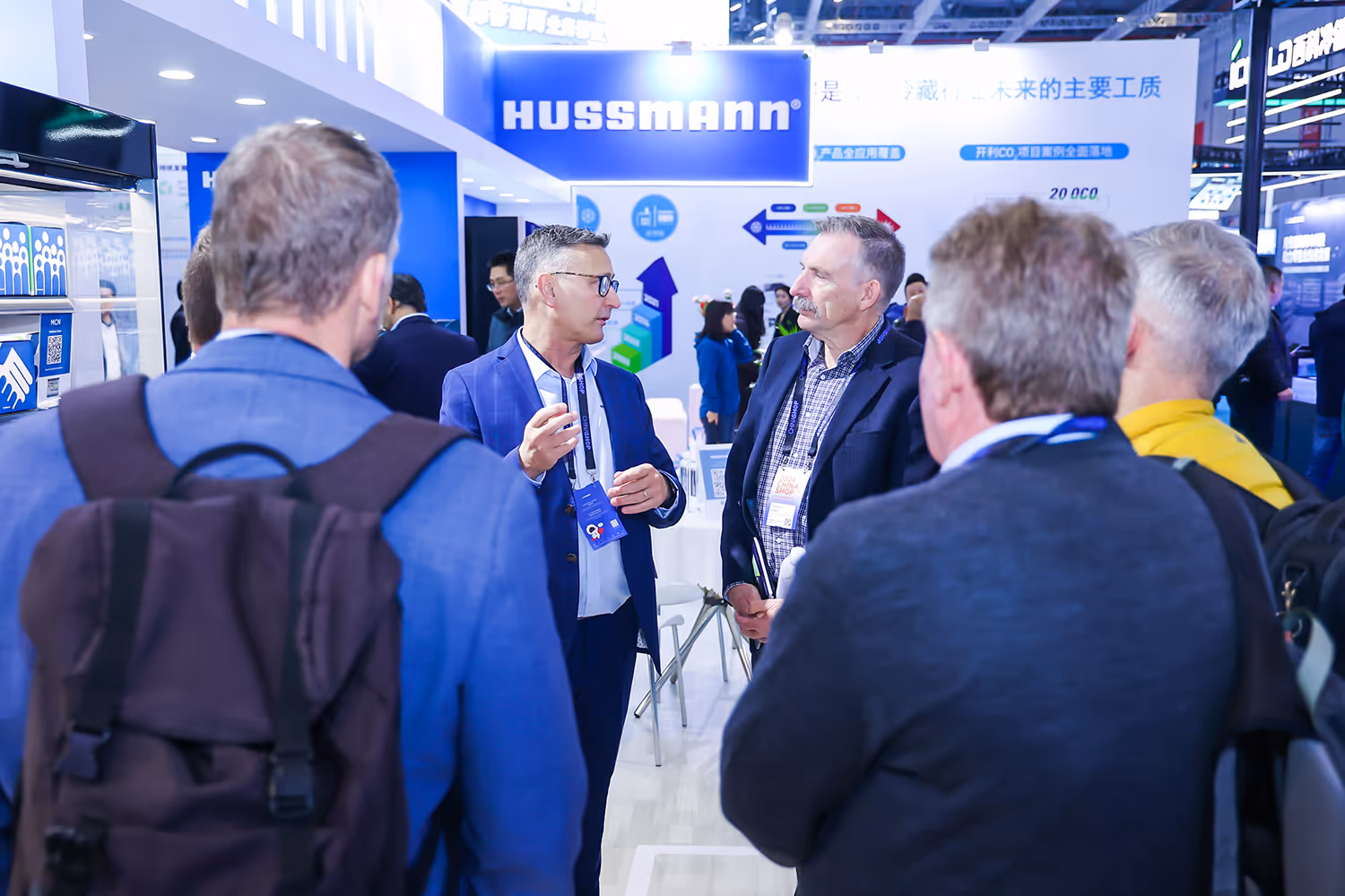 Two men in business attire talking at a Husmann exhibition booth with several people around.