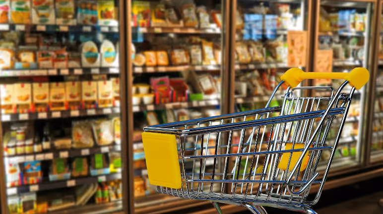 Empty shopping cart with yellow handle in front of grocery freezer shelves filled with various packaged food products.