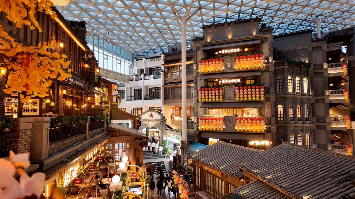 Indoor shopping street with traditional-style buildings, colorful lanterns, warm lighting, and glass roof ceiling.